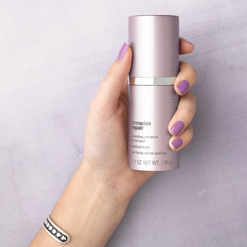 Hand with nails painted purple holding bottle of TimeWise Repair Revealing Radiance Facial Peel from Mary Kay against purple background.