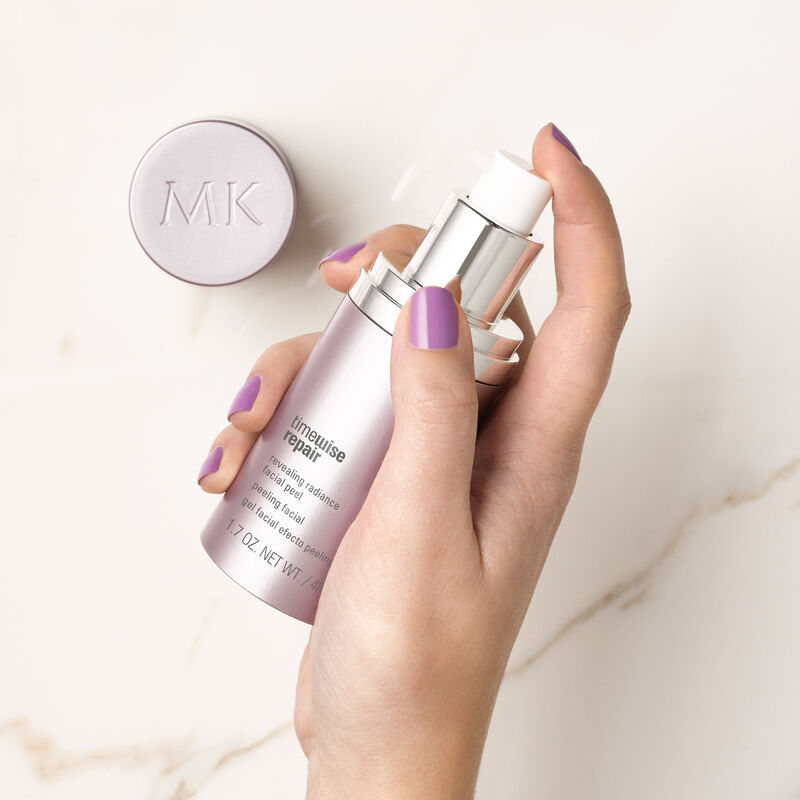 Hand with nails painted purple holding bottle of TimeWise Repair Revealing Radiance Facial Peel from Mary Kay with cap on marble counter.
