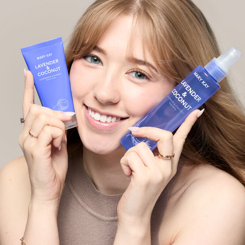 A white model with blue eyes holds the lavender and coconut hand cream and body spray by her face while smiling.&nbsp; image number null