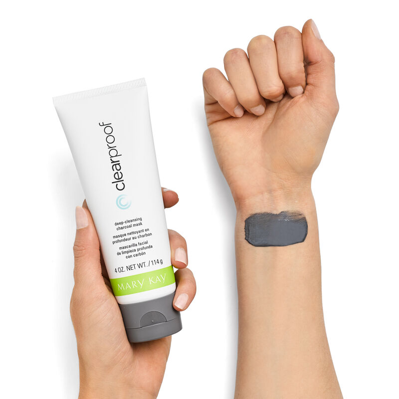 A model holds the Mary Kay Clear Proof Deep Cleansing Charcoal Mask bottle in her left hand while a smear of the grey mask appears on her right wrist 