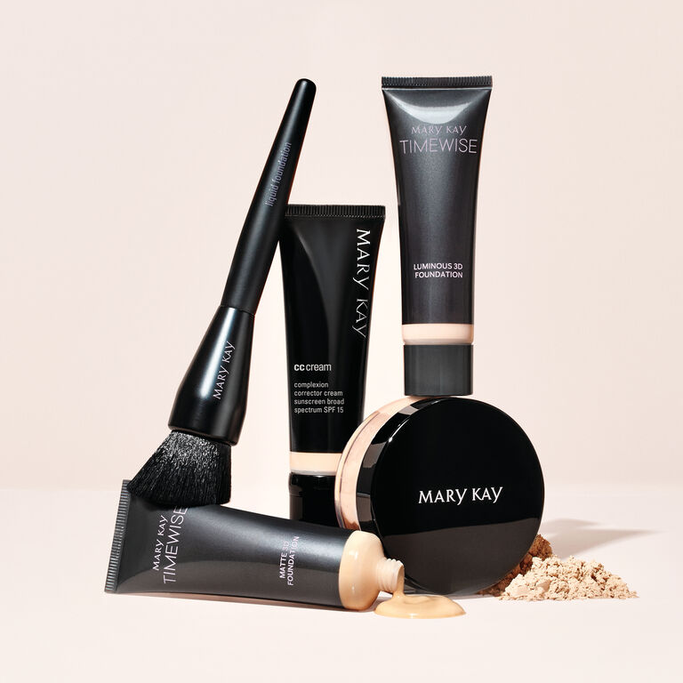 Mary Kay TimeWise foundation, CC cream, and makeup brush arranged with loose powder on a light background.